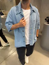 Camisa de jean de corte boxy, color azul claro, con mangas cortas, cuello clásico y cierre frontal con botones metálicos. Presenta un bolsillo frontal en el pecho.