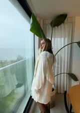 Vestido corto color crudo con diseño holgado, cuello redondo y mangas largas con puños acampanados. Presenta detalles de encaje floral en el cuello y en los puños de las mangas.