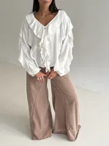 Blusa blanca de corte holgado con escote en V, decorada con volados frontales y en las mangas. Presenta mangas largas con puños elásticos y cierre frontal con botones.