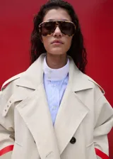 Chaqueta larga de algodón color beige, de diseño recto con cuello en pico y solapas con muesca. Cuenta con cierre delantero de doble botonadura, bolsillos laterales y mangas largas reversibles.