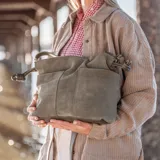 Cartera tipo bucket de cuero gamuzado color verde oliva, con correa de hombro y correa bandolera ajustable. Tiene dos bolsillos externos, cierre con broche imán y forro interno de gabardina de algodón con tres compartimentos.