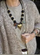 Collar de diseño artesanal con cuentas esféricas negras y separadores dorados. Presenta un conjunto de dijes colgantes que incluyen un corazón dorado texturizado, una cruz con detalles en azul, un dije de ojo turco, una cuenta rectangular marrón y una cuenta de piedra natural azul.