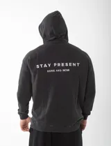 Canguro con capucha de felpa premium, color negro con efecto lavado a la piedra. Presenta un diseño oversize unisex con la frase 'STAY PRESENT HERE AND NOW' bordada en la espalda.