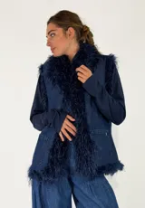 Chaleco de denim azul con detalles de flecos de pelo sintético en los bordes, cuello y solapas. Presenta bolsillos frontales tipo parche.