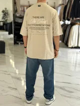 Remera oversize color beige, con mangas cortas y cuello redondo. Presenta un estampado tipográfico en la espalda con la frase "There are no facts, only interpretations" y otros detalles de diseño.