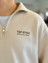 Buzo marrón de felpa con medio cierre y cuello alto, manga larga y puños elásticos. Presenta el texto "TOP STYLE follow your dreams" bordado en el pecho.