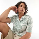 Camisa de manga corta con estampado de hojas en tonos verdes y blancos, cuello tipo campana y cierre frontal de botones.