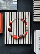 Collar corto de diseño artesanal compuesto por cuentas de diversas formas, texturas y materiales en tonos rojo, marrón, beige y blanco, con cadena de ajuste metálica.