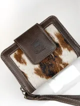 Mini billetera de cuero marrón con detalles en cuero con pelo con estampado animal print tipo vaca en tonos blancos, marrones y negros. Cuenta con solapa frontal con botón a presión y diseño de búho grabado, ribetes pespunteados y correa de mano.