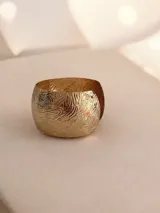 Brazalete tipo cuff de metal dorado con diseño de relieve ondulado y acabado brillante. Es de diseño abierto y ajustable.