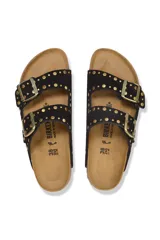 Sandalias Birkenstock modelo Arizona con dos tiras ajustables de gamuza negra decoradas con remaches metálicos dorados. Presenta hebillas doradas, plantilla anatómica de corcho y suela de goma negra.
