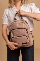 Mochila urbana de diseño funcional en color beige, confeccionada en tela impermeable. Cuenta con un compartimento principal amplio, un bolsillo secundario y un bolsillo frontal, todos con cierre. Incluye asa superior y correas ajustables para los hombros, con detalle de etiqueta de marca en el frente.