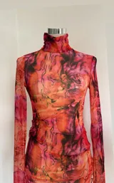 Vestido corto de microtul con estampado abstracto en tonos naranja y fucsia. Tiene cuello alto, mangas largas acampanadas y abertura lateral en la falda.