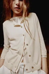 Chaqueta efecto doble de punto color beige con cuello redondo, manga por debajo del codo y cierre frontal con botones. Detalle de cuello y bajo de camisa interior asomados.