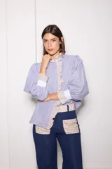 Camisa de manga larga con diseño combinado en tres telas, presentando un patrón de rayas verticales en azul y blanco. Cuenta con cuello alto, volados frontales y puños en contraste. El diseño es holgado con mangas abullonadas.
