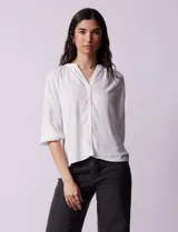 Blusa blanca de gasa marca Sioni, con cuello a la base y escote en V. Tiene abertura central con botones y mangas 3/4 con terminación fruncida.