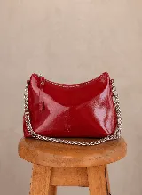 Cartera tipo media luna de cuero charolado en color rojo, con cierre superior y correa de cadena metálica plateada. Presenta el logo de la marca grabado en la parte inferior frontal.