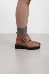 Botines de cuero vacuno color suela con textura envejecida, suela track negra con pespunte en contraste, solapa cruzada con gran hebilla metálica plateada y cierre lateral interior.