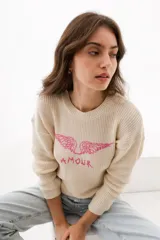 Sweater de punto color beige, con cuello redondo y mangas largas. Presenta un diseño bordado en el frente con alas y la palabra 'AMOUR' en color fucsia.