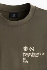 Remera de algodón de punto pesado con efecto lavado, color verde militar. Presenta un corte boxy (holgado) con cuello redondo ribeteado y un pequeño estampado blanco en el pecho con texto y logos.