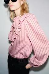 Camisa de manga larga con cuello subido, cierre frontal con botones y detalle de volantes en el pecho. Presenta un diseño a rayas verticales y puños elásticos.