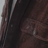 Campera color borgoña de pana con cuello camisero, cierre frontal con botones a presión y dos bolsillos de parche en el pecho. Interior acolchado con patrón de rombos.