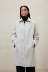 Chaqueta larga color beige, con cuello camisero y cierre frontal de botones a presión. Tiene dos bolsillos laterales y está confeccionada con poliamida y poliéster 100% reciclados.