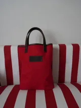 Matera de lona color rojo con mangos de cuero marrón. Presenta un diseño rectangular con base reforzada y parche frontal de cuero con logo grabado.