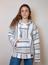 Buzo estilo mexicano con capucha, de tejido grueso a rayas horizontales en tonos blanco, beige y negro. Tiene bolsillo tipo canguro y cordones ajustables en la capucha.