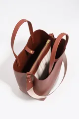 Bolso de hombro de tamaño mediano color marrón, confeccionado en piel vacuna. Presenta doble asa corta de mano y correa de tejido jaspeada con el nombre de la marca estampado. Incluye detalles en piel, cierre con botón de presión, logo grabado en dorado en el frente y acabados metálicos.