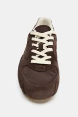 Zapatilla de running en combinación de materiales, con cuerpo de piel y detalles en gamuza, color marrón. Cuenta con cierre mediante cordones en tono crema y suela plana de goma.