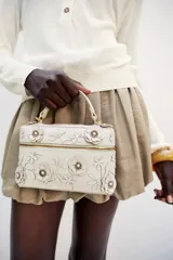 Bolso de mano formato vanity color crudo, con apliques de flores en relieve y hojas en el cuerpo. Cuenta con asa corta superior y cierre con cremallera doble dorada.