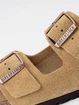 Sandalias Birkenstock modelo Arizona, confeccionadas en gamuza color tostado. Presentan dos correas ajustables con hebillas metálicas grabadas, plantilla anatómica de corcho y látex, y suela de goma.