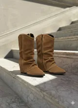 Botas de caña media confeccionadas en gamuza color marrón, con diseño slouchy y cierre lateral. Presentan una silueta de punta fina y taco cuña oculto.