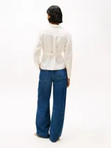 Blusa blanca de lino con diseño cruzado, cuello camisero, mangas largas con puños abotonados, dos bolsillos con solapa en el pecho y cinturón anudable.