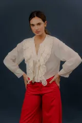 Camisa de algodón color crudo con volados delanteros y detalles de puntilla bordada. Presenta escote en V con botones y mangas largas abullonadas con puños ajustados.