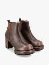 Bota corta tipo botineta de cuero negro con taco cuadrado y plataforma redondeada. Presenta un panel elástico lateral en color crema y tirador en la parte trasera.