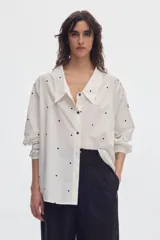 Camisa oversize blanca con estampa de lunares negros, cuello en pico con solapas clásicas y mangas abullonadas con puños elásticos.