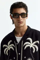 Camisa de manga corta de H&M, con corte relajado, cuello abierto estilo resort, tapeta frontal con botones y bajo recto. Diseño estampado en color blanco sobre base negra con motivos de palmeras, soles y estrellas.
