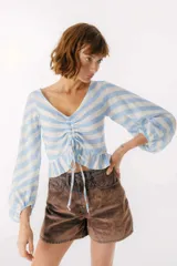 Top de lino italiano a rayas horizontales celestes y blancas, con escote en V ajustable mediante jareta central con lazos, mangas largas estilo globo y volado en el ruedo inferior.