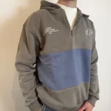 Canguro con capucha y cierre frontal parcial, de diseño color block en azul grisáceo y blanco. Presenta bordado con logo y texto en el pecho.