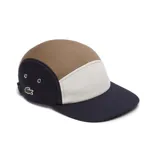 Gorra de cinco paneles Lacoste con diseño tricolor en tonos azul marino, beige y blanco. Presenta el logo del cocodrilo bordado en el lateral y ojales metálicos para ventilación.
