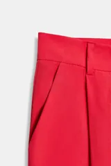 Pantalón de tiro alto color rojo vibrante, de corte ancho y pierna recta, con pliegues marcados en la parte delantera. Presenta cierre frontal con botón y cremallera, y bolsillos delanteros.
