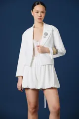 Saco corto de color blanco con solapas clásicas, mangas largas y detalle de parche rectangular con texto en el puño izquierdo. Presenta una silueta estructurada y elegante.