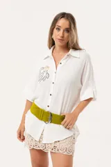 Camisa blanca de silueta oversize, con cuello camisero, manga 3/4 y corte más largo en la espalda. Presenta bordados de coral y caracol en el frente, y botones a tono.