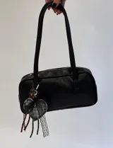 Cartera tipo shoulder bag de cuero sintético negro, con cierre superior y doble asa de hombro. Incluye un llavero colgante con forma de corazón acolchado en tela a cuadros, adornado con encaje y cintas colgantes.