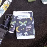 Billetera elástica de tejido resistente, color negro con estampado de mariposas, lunas, hongos y estrellas, con anilla lateral para llavero.