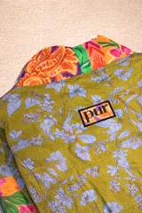 Bata tipo kimono de diseño patchwork, confeccionada con una mezcla de telas estampadas con motivos florales, paisley y geométricos en tonos verdes, naranjas y violetas. Presenta mangas tres cuartos, aberturas laterales en el ruedo y un lazo ajustable en la cintura.