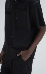 Camisa manga corta de estilo técnico, color negro, confeccionada en microfibra liviana. Presenta cuello camisero, cierre frontal con broches a presión ocultos y doble bolsillo frontal utilitario con detalles de red y cierre.