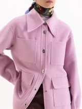 Chaqueta de paño color rosa, con cuello camisero, cierre frontal con botones metálicos y cuatro bolsillos plaqué delanteros. Presenta mangas largas y costuras con pespuntes decorativos al tono.